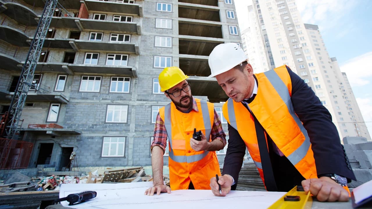 Two construction supervisors in hard hats and orange vests reviewing site plans together