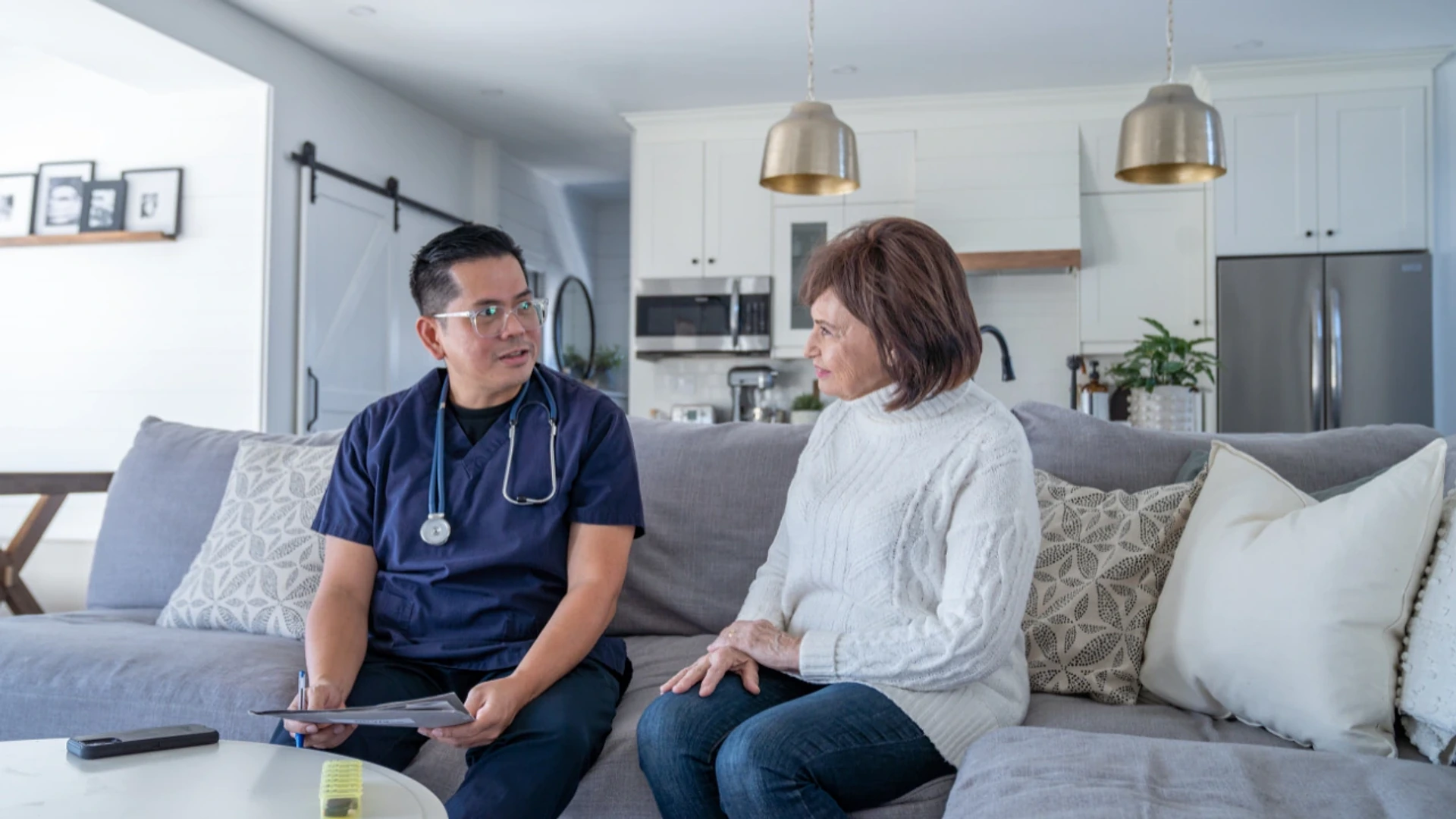 A home care nurse in scrubs talking with an older woman on the sofa in her living room
