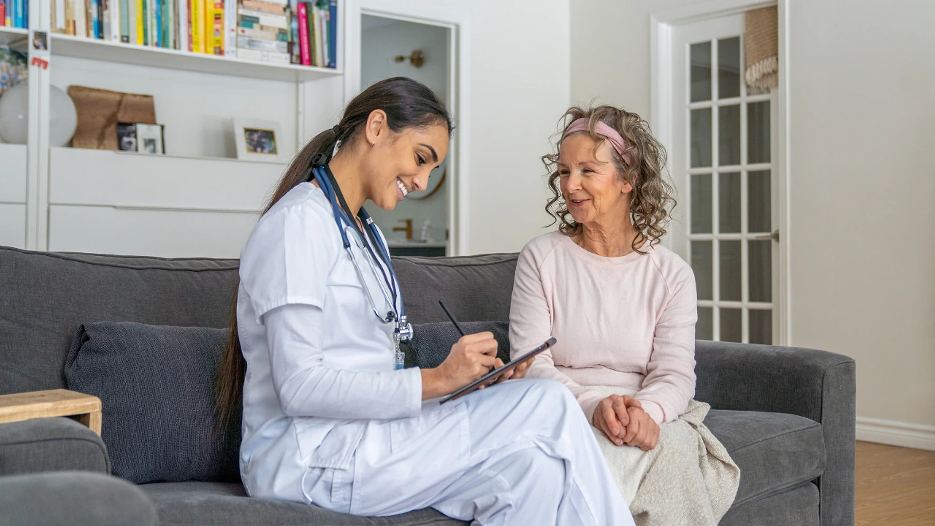 A home health nurse filling in a form on a tablet while talking with an older woman at home