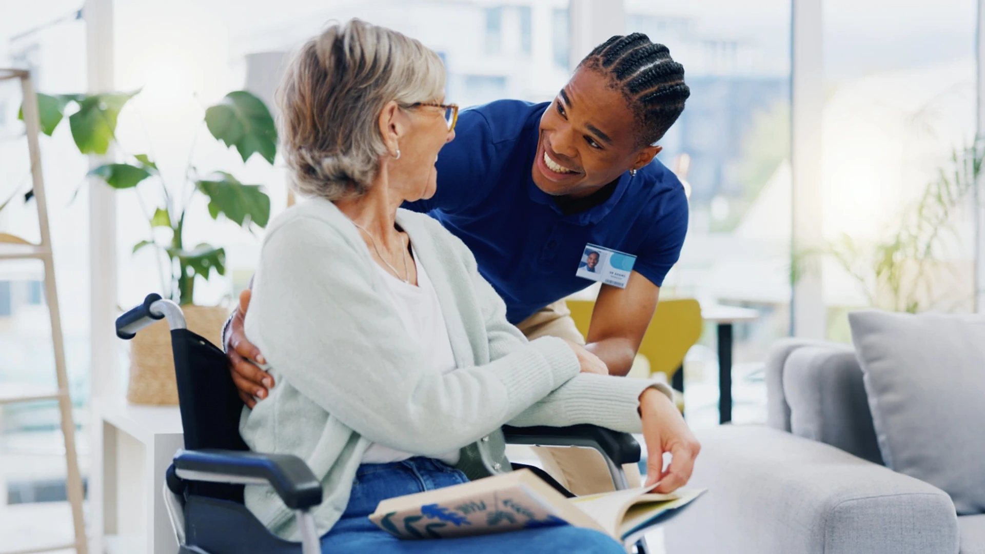 A smiling caregiver with a name badge leaning in to talk with an older woman in a wheelchair