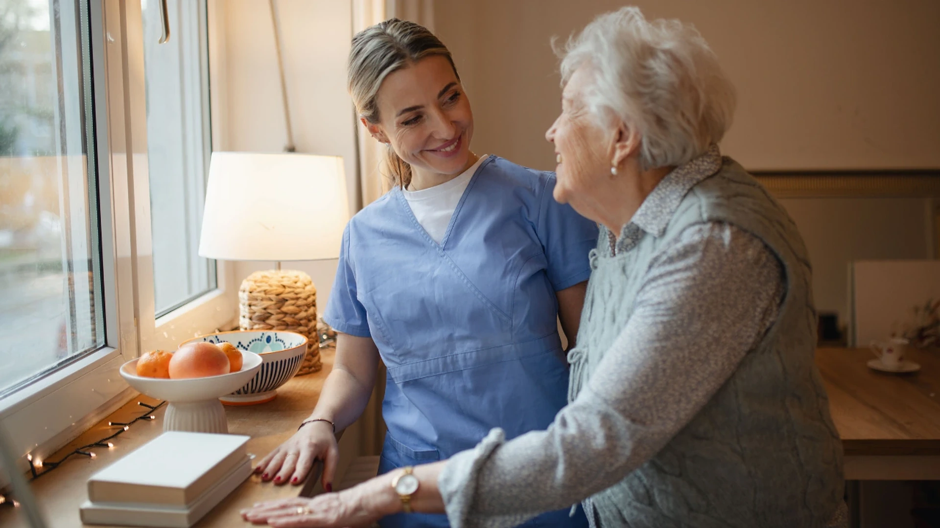 A caregiver in scrubs smiling at an older woman by a window at home
