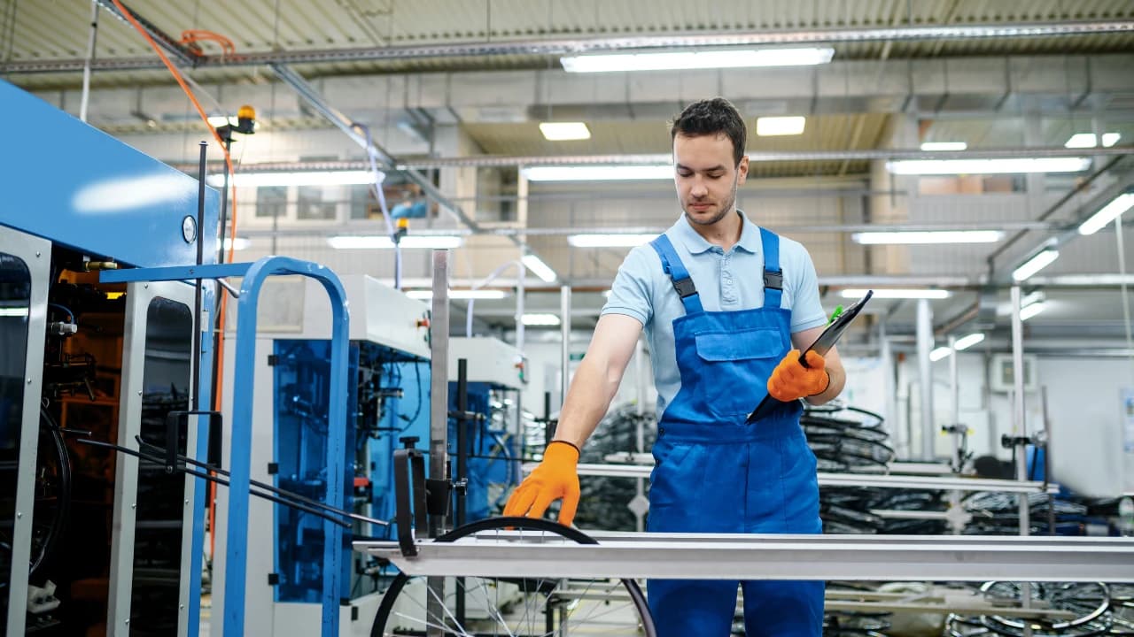 A production worker in blue overalls with a clipboard on an assembly line