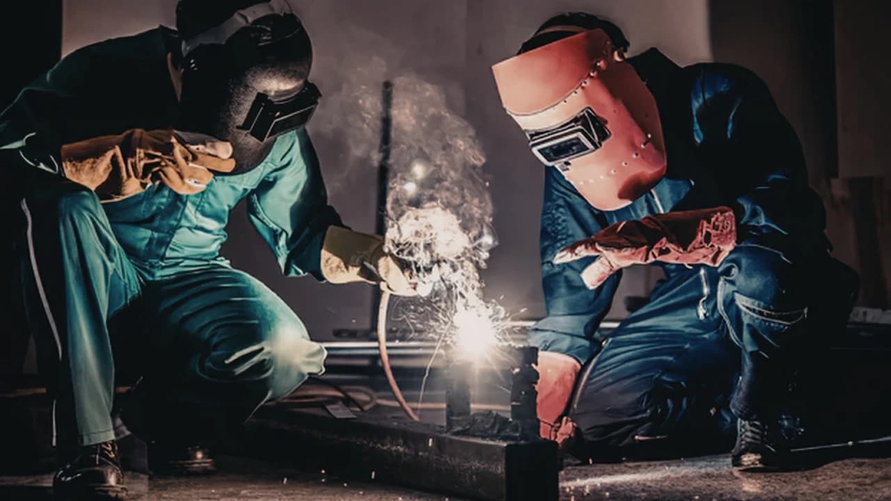 Two welders in protective gear working on a steel part with sparks flying