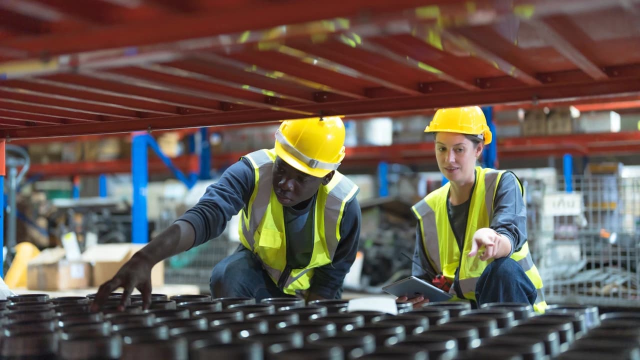 Two warehouse workers in hard hats and high-visibility vests checking a tablet among stock